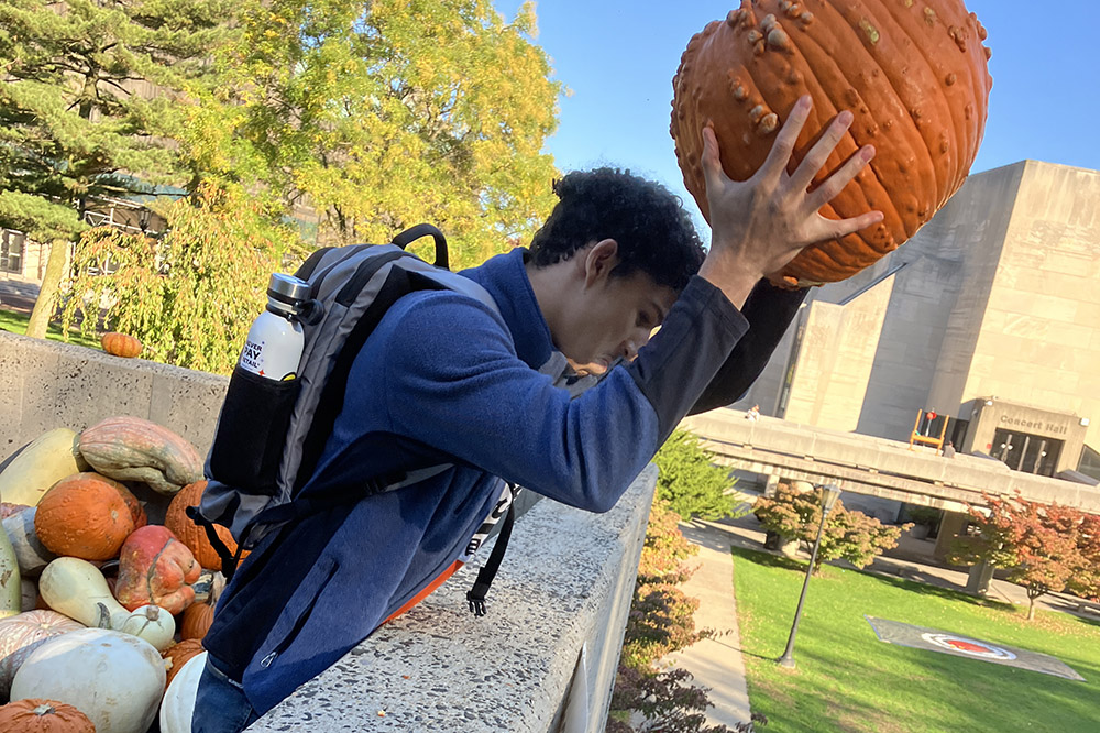 A person in a backpack and blue shirt drops an orange pumpkin off a rooftop