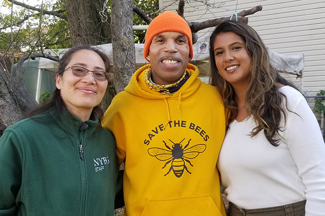 Three people—one in a green sweatshirt, one in a yellow sweatshirt, and the third in a white long-sleeve shirt—pose together for an outdoor photo