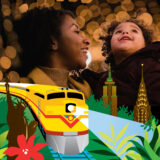 A parent and child enjoy an illuminated holiday scene as a yellow locomotive passes them on a track