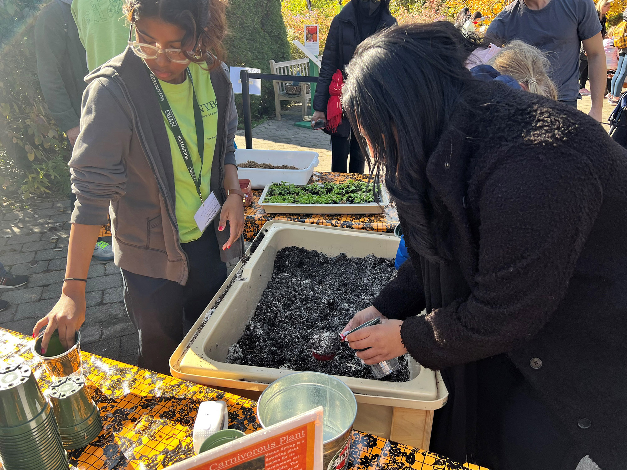 A person in a black coat packs soil from a large bin as another person in a gray sweatshirt and green shirt helps