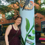 A person in a purple dress leans against a pillar decorated with caterpillars and orange butterflies