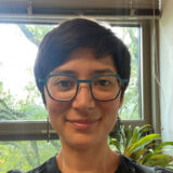 A person with short dark hair and blue glasses poses for a photo in a room with plants