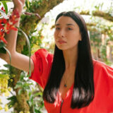 Person standing with long brown hair wearing red shirt and arm up along tropical archways with red orchid flowers and other orchids along the arches.
