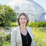 Head shot of Jennifer Bernstein, wearing a black v-neck shirt with a tan blazer and silver and gold long lariat necklace. Gold round earrings and short blond hair, with an upturned mouth, and blue eyes. Standing in front of a garden with yellow and purple flowers that are blurry, and a green hedge and the glass dome of the Enid A. Haupt Conservatory.