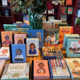 Multiple colorful books of African American children on the cover and other brightly colored covers of books on a table, with a vase and green plants coming out the top.