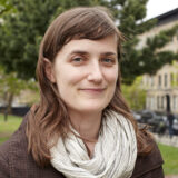 A person with long brown hair with bangs wearing a cream colored infinity scarf and purple sweater looks at the camera