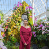 Lily Kwong standing in a pink jumpsuit in front of a mountain installation adorned with orange, purple, and white orchids