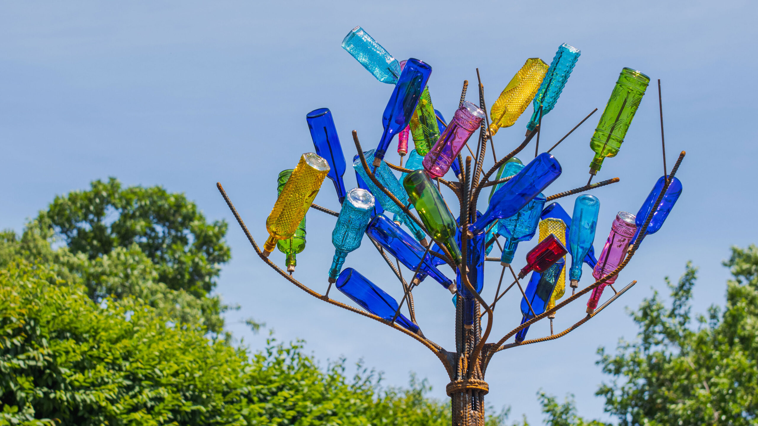 Blue, yellow, green, and pink translucent glass bottles upside-down on a metal tree branch structure against a blue sky background with green bushes along the bottom view.