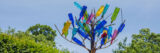 Blue, yellow, green, and pink translucent glass bottles upside-down on a metal tree branch structure against a blue sky background with green bushes along the bottom view.