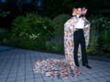 A person in black pants, a white shirt, and a long, flowing floral cape poses for a photo after dark