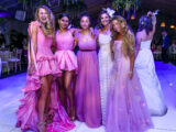 Six people in pink gowns pose for a photo on a dance floor