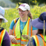 A group of people in safety vests meet in a green field to examine plants