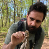 A person with a dark beard and short dark hair examines insects in the forest