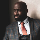 A person with a short black beard, wearing a gray suit and orange tie, poses for a photo