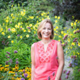 A woman with short blonde hair and a pink dress stands in front of yellow and purple flowers and lots of green foliage