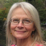 a close up of a person with shoulder length white hair, wire glasses in a pink top