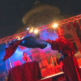 a DJ on stage with a performer twirling a fabric flag under red stage lights