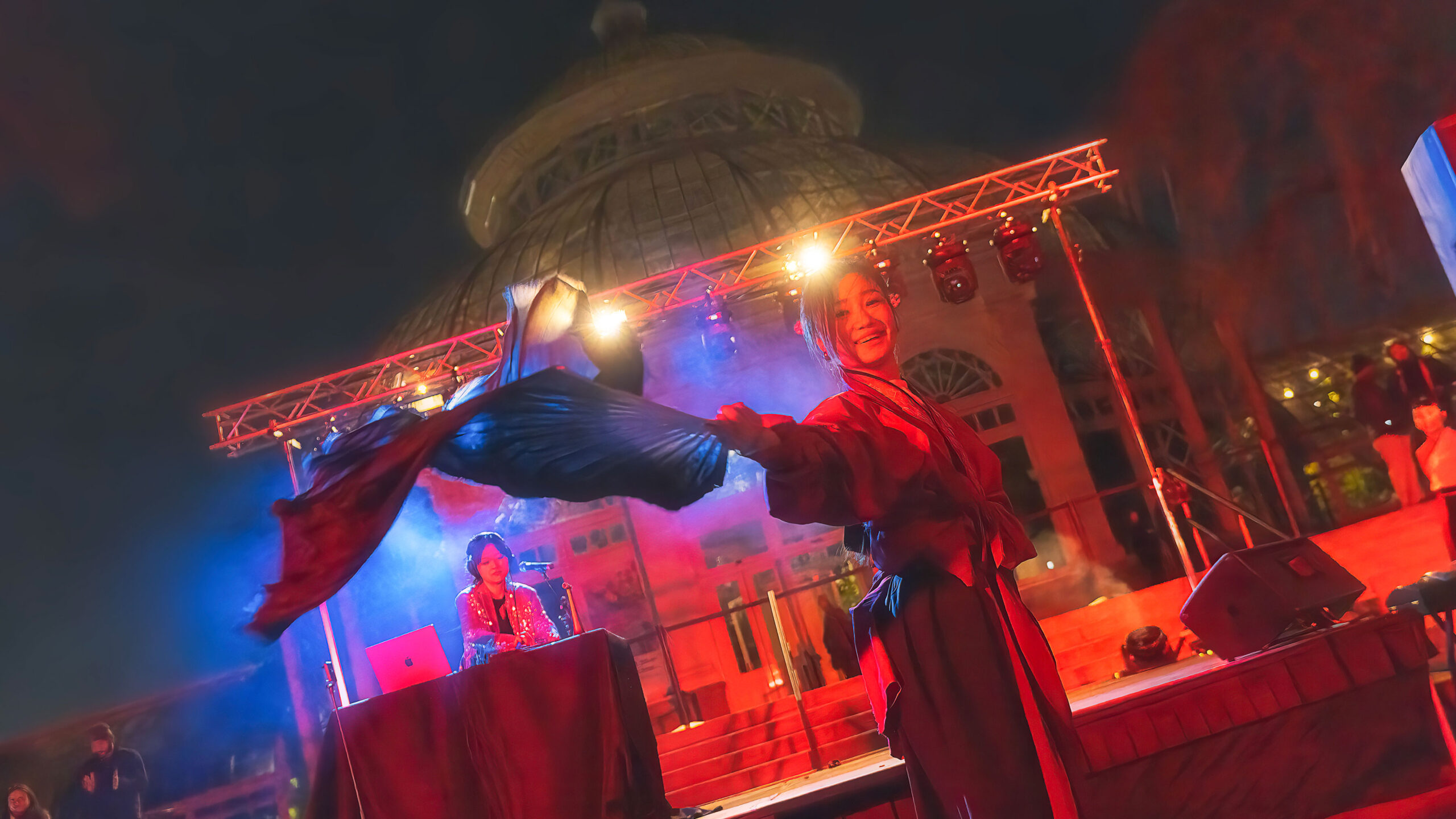 a DJ on stage with a performer twirling a fabric flag under red stage lights