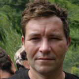 A man with short brown hair and a black shirt looks directly at the camera with a soft smile.