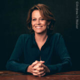 An image of actress Sigourney Weaver, who smiles wide with her hands clasped and resting on the table in front of her.