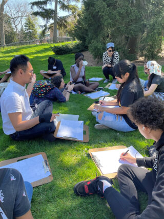 Students take notes while gathered in a group, sitting on the grass outdoors