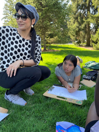 A student and teacher take notes while engaged in an outdoor workshop on a green lawn