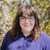 A person in a purple shirt and glasses with long brown hair poses for a photo outdoors