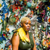 Person with short cut blonde hair and large yellow feather earring, looking out at the audience. Wearing olive green leather jump suit. Sitting in front of multi-colored, and textured wall art with tiger print, flowers, and many other brightly colored fabrics and patterns.