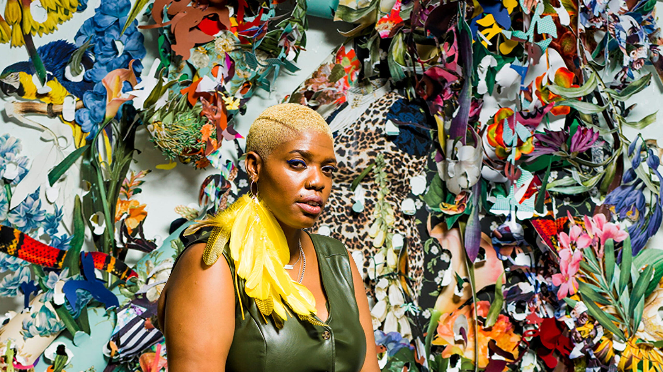 Person with short cut blonde hair and large yellow feather earring, looking out at the audience. Wearing olive green leather jump suit. Sitting in front of multi-colored, and textured wall art with tiger print, flowers, and many other brightly colored fabrics and patterns.