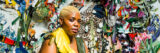 Person with short cut blonde hair and large yellow feather earring, looking out at the audience. Wearing olive green leather jump suit. Sitting in front of multi-colored, and textured wall art with tiger print, flowers, and many other brightly colored fabrics and patterns.