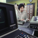 A person in an antique photo examines library materials in a room full of cabinets and computers