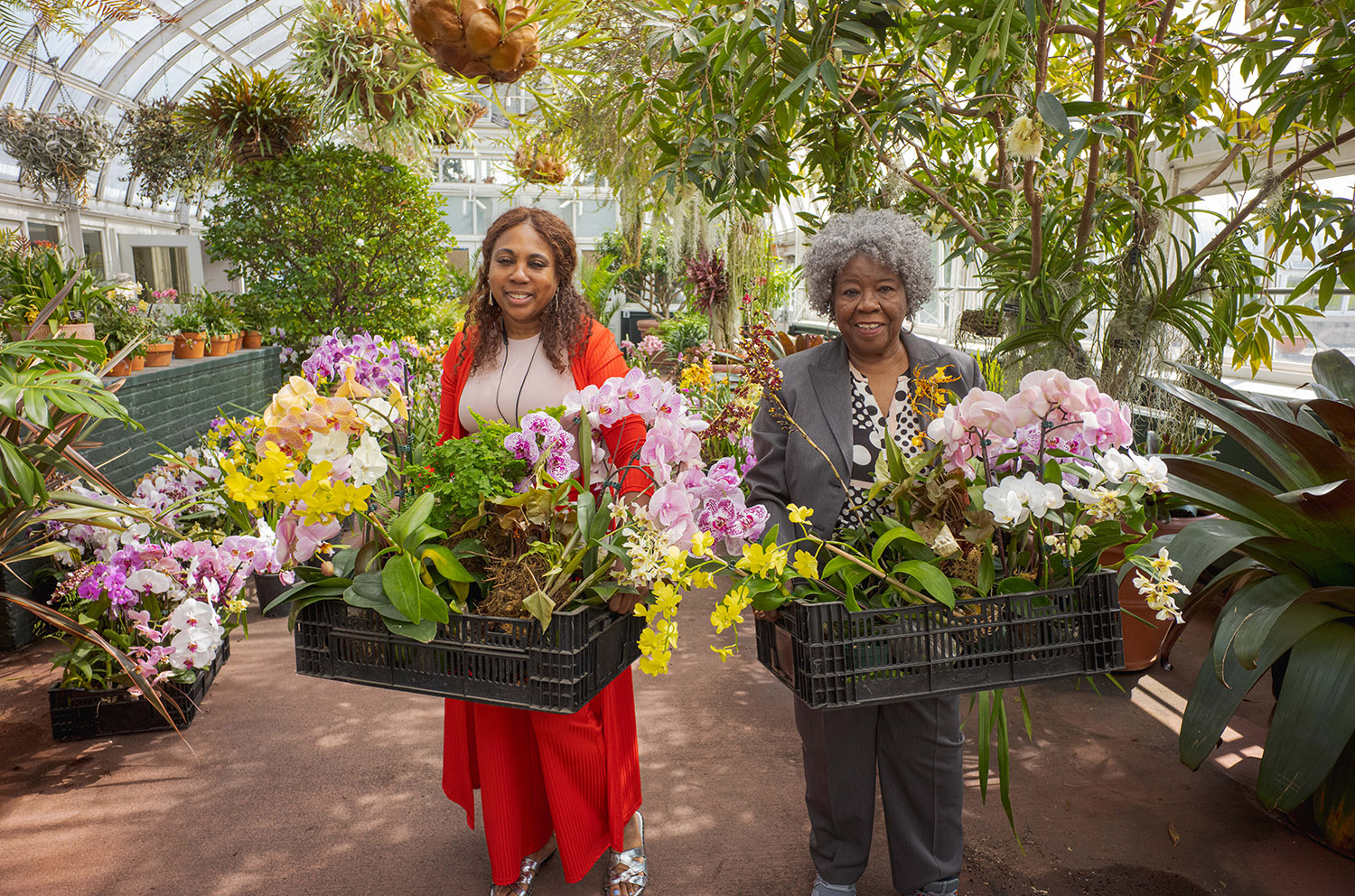 Two people in red and black outfits carry baskets of colorful flowers through a conservatory