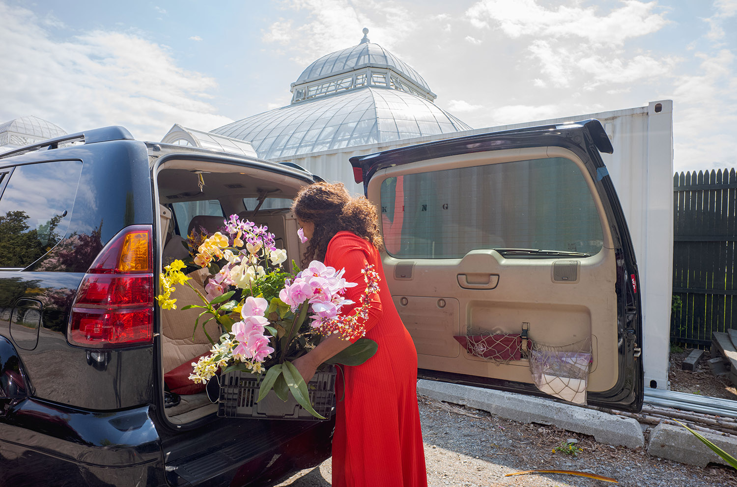 A person in a red dress packs pink and yellow orchids into the back of a car on a sunny day