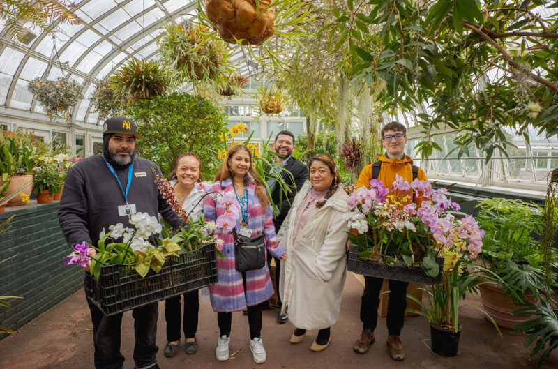 A group of people in a verdant greenhouse carries baskets of colorful flowers