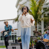 A person in blue pants and a flowing white shirt sings into a microphone as a drummer and guitarist back up their performance