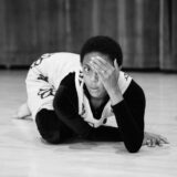 a person in black and white clothing crouched on the floor with one hand over their eye