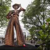 A person standing in a pose with their hand holding a hat in a brown and gold pants and shirt outfit at the end of a black runway in front of the glass conservatory dome.