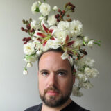 Man wearing elaborate white floral headpiece, with various size flowers and some smaller darker flowers in the edges. Sitting against a stark grey background.
