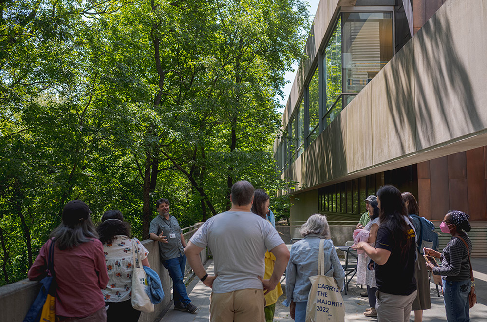 People gather around a tour guide on a sunny spring day outside of a tan building, bordered by green trees