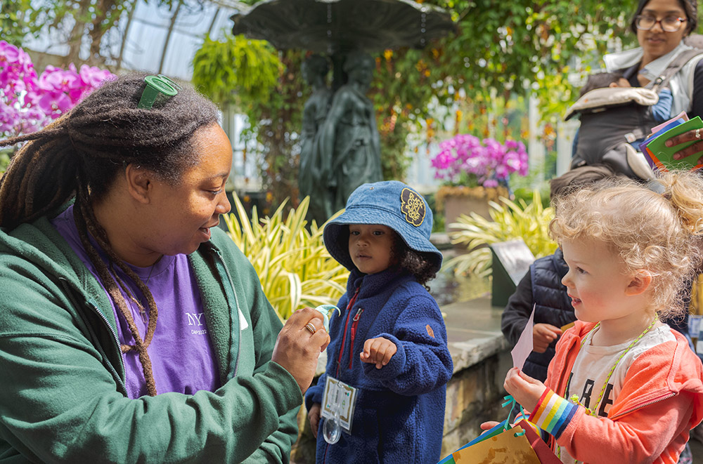 A person in a purple shirt and green hooded sweatshirt shows a plant to a group of small children in a conservatory.