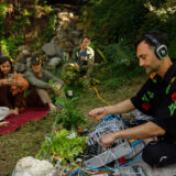 a person with headphones on sits in a wooded area with equipment in front of them connecting to the plants in the area