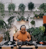 Headshot of DJ Niyah West sitting in front of DJ equipment wearing a sleeveless dark yellow top and headphones. In the background are many mounted green plants along a white wall.