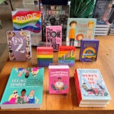 Collection of colorful books on Pride, and other LGBTQ+ topics, ranging for adults and kids on a brown wooden table.