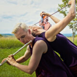 Two people in purple tops pose with instruments in a field. One person leans playing on a violin while the other plays the flute.