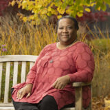 Person sitting on a bench wearing a long sleeve patterned dark pink and red shirt with black leggings. Behind them are tall grasses and yellowing leaves with some green still on a tree.