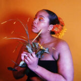 a person with orange flowers in a dark-haired bun, in a black tube top holding grain and green plants in their hands in front of an orange background