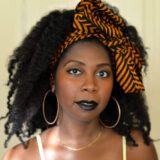 Head shot of person wearing an orange and black striped fabric head band, and large gold hoop earrings, with skinny white tank top sleeves.