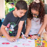 an adult with a white apron and glasses with a child in a graphic t-shirt color in a canvas bag on a yellow and flower designed table