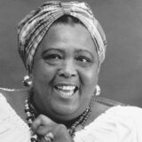 Black and white photo of person with patterned fabric headpiece tied on top of their head. Wearing hoop metal earrings and light colored ruffled top with upturned lips.