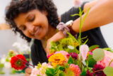 a close-up photograph of a person with short curly hair arranges a flower in a pink and orange bouquet